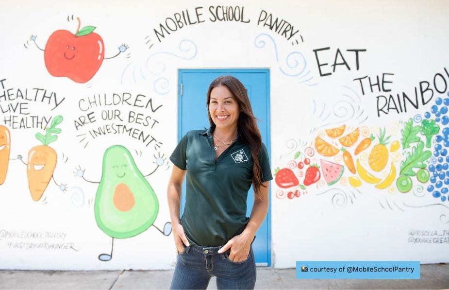 Zeina Zein-Wolland smiling in front of a mural featuring fruits, vegetables, and the words “Mobile School Pantry – Eat the Rainbow.”